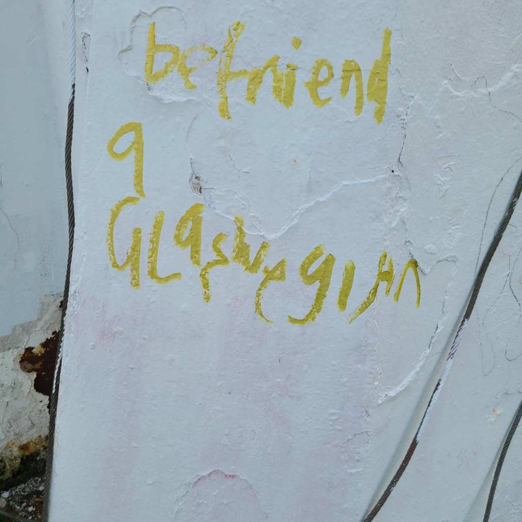Yellow graffiti on a white flagpole, which says: befriend a Glaswegian