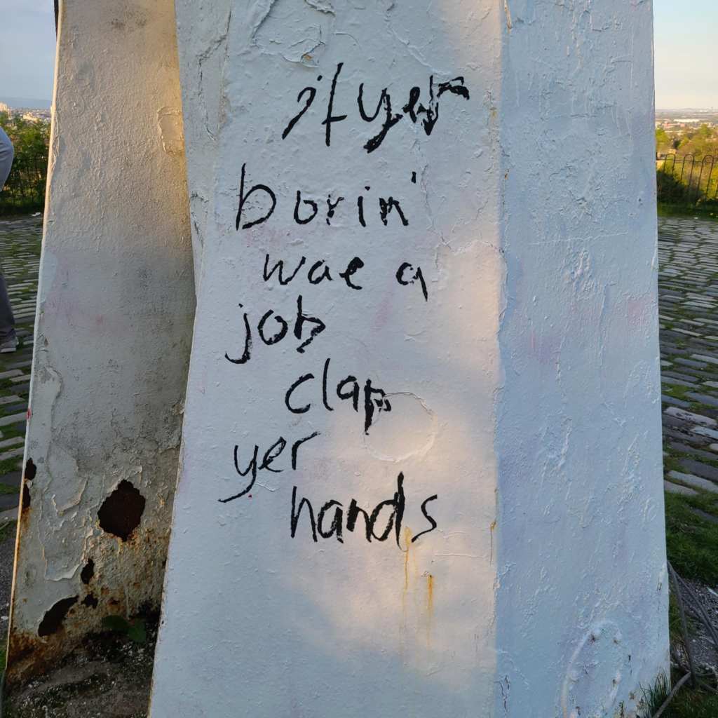 Black graffiti on a white flagpole, which reads: if yer borin' wae a job clap yer hands